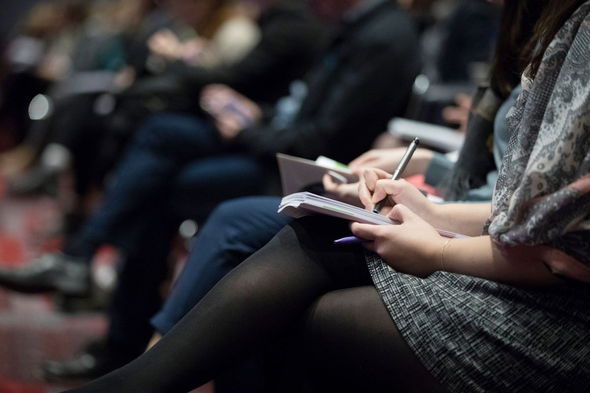 participation-presentations-congres-2018 Public assis en rangées lors d'une conférence, personnes prenant des notes sur un carnet, dans une salle aux lumières tamisées.