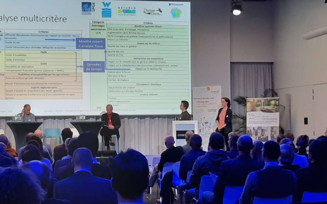 Conférence en salle avec projection d'un tableau d'analyse multicritère à l'écran, trois intervenants sur scène face à un public nombreux assis.