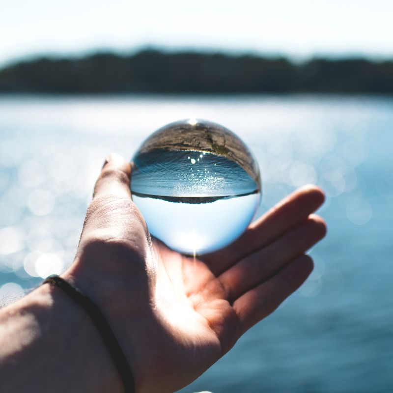 Main tenant une boule de cristal au-dessus d'un lac, reflétant en miniature le paysage de collines boisées inversé, sur fond de rives embrumées.